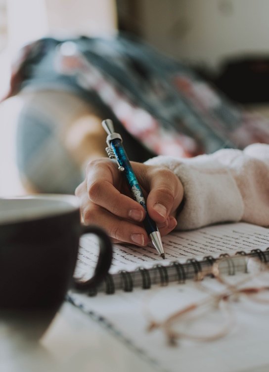 Iemand schrijft in een notitieboekje met een blauwe pen. Er staan een kopje en een glas bij de hand.