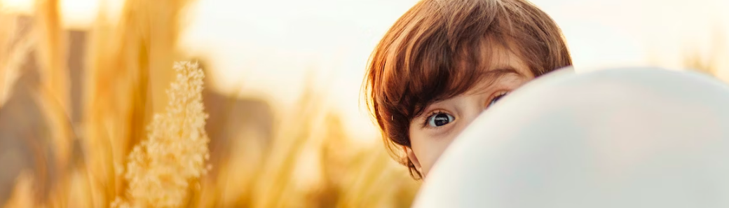 Een kind is gedeeltelijk verborgen achter een witte ballon in een veld met hoog gras en kijkt recht in de camera.