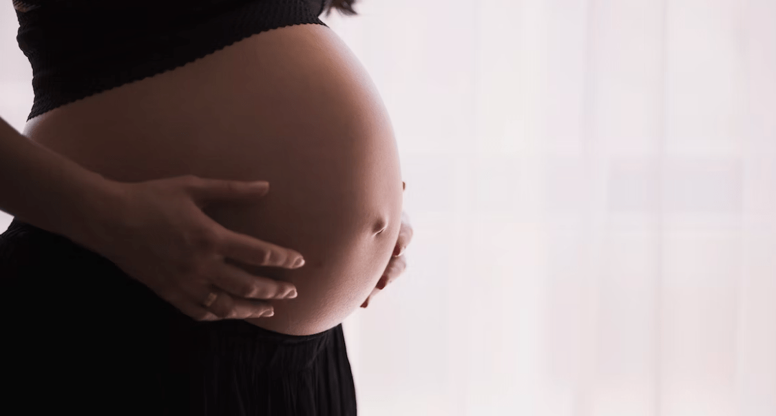 Een close-up van een zwangere vrouw die haar buik voorzichtig vasthoudt, gekleed in een zwart topje, voor een zacht verlichte achtergrond.