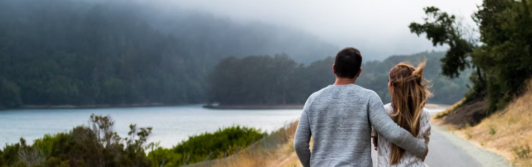 Een stel wandelt over een geplaveid pad langs een rustig meer, omgeven door in mist gehulde heuvels en bomen.