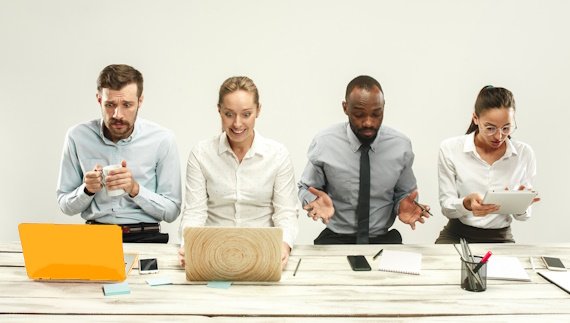 Vier mensen zitten aan een tafel met laptops en tablets. Ze hebben een gemengde, geconcentreerde en verwarde blik.