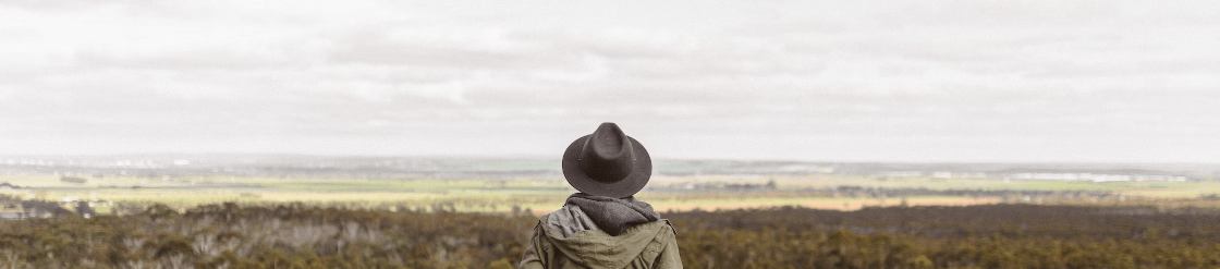 Een persoon met een hoed staat op een heuveltop en kijkt uit over een uitgestrekt landschap van velden en de verre horizon onder een bewolkte hemel.