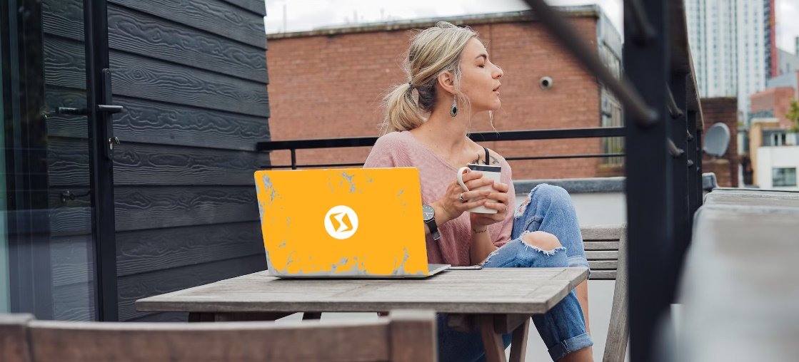 Een vrouw zit aan een tafeltje op het terras, heeft een laptop, een mok in haar hand en haar ogen gesloten.