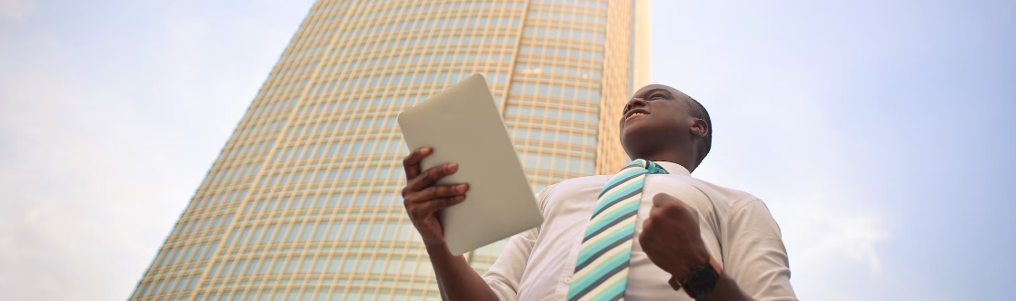 Een persoon staat voor een hoog gebouw, houdt een tablet vast en balt een vuist. Hij lijkt gemotiveerd of triomfantelijk.